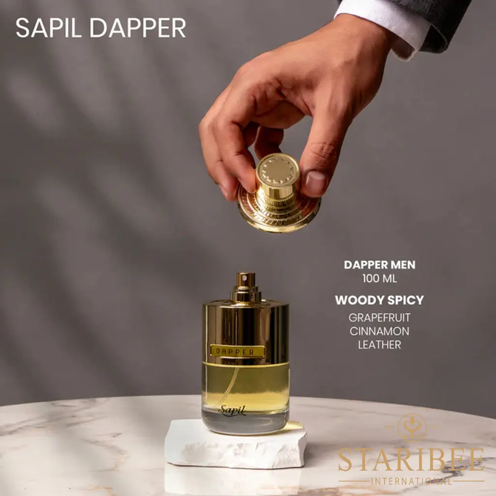 Hand holding a gold cap above a perfume bottle labeled 'Sapil Dapper' on a marble surface with a gray background.