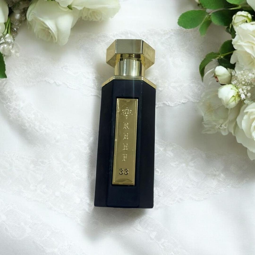 Reef 33 luxury perfume bottle with black and gold design placed on a white lace background surrounded by white flowers, highlighting an elegant floral woody fragrance aesthetic.